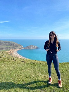 A white female with long brown hair smiles at the camera on a coastal path. 